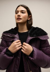 Lilla puffjakke musta fliiski krae ja lukuga. Sile tekstuur, laiad varrukad ja kullakarva detailid.