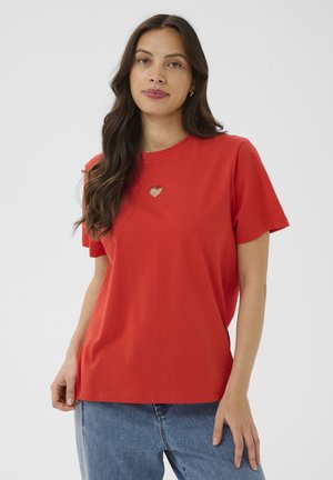 Vrouw met lang donker haar, gekleed in een rood T-shirt met een kleine hartvormige uitsparing bij de halslijn en een blauwe spijkerbroek.