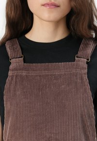 Avestito in velluto a coste marrone, caratterizzato da righe testurizzate e spalline regolabili con fermagli in metallo. Indossato con una maglietta nera sotto.
