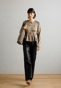 Beige long-sleeve blouse with button front and gathered waist, paired with black wide-leg pants and patterned sneakers. Neutral backdrop.