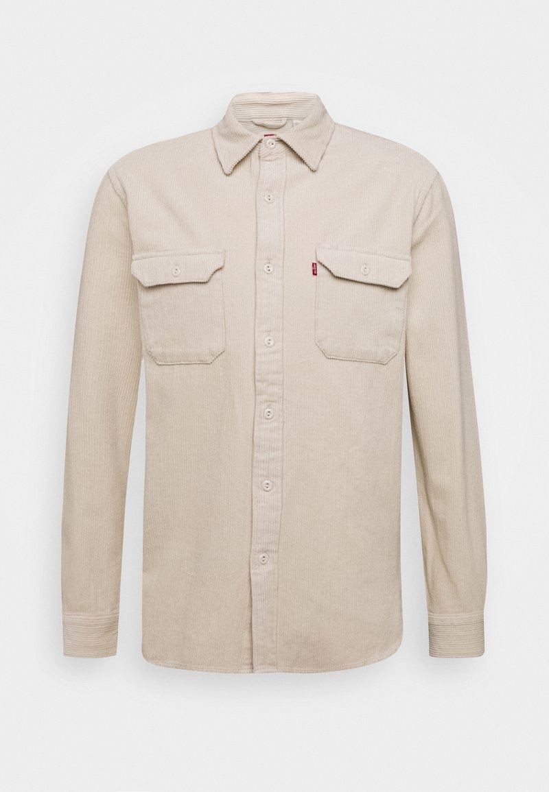 Chemise beige en velours côtelé à manches longues avec boutons devant et deux poches poitrine boutonnées sur un fond uni.