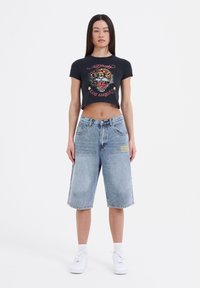 Sort cropped t-shirt med grafik af en tiger og tekst, parret med lyseblå denim bermudashorts med en gul designdetalje. Hvide sneakers.