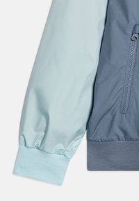 Vaaleansininen ja tummansininen kevyt takki, jossa on vetoketju, resorit hihansuuissa ja sileä pinta. Takissa on kontrastivärinen blokkimainen muotoilu.