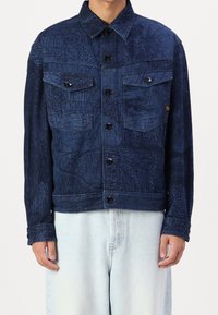 Mörkblå denimjacka med ett strukturerat mönster, med två bröstfickor, knäppning med knappar och rak passform, som matchas med ljusa jeans.