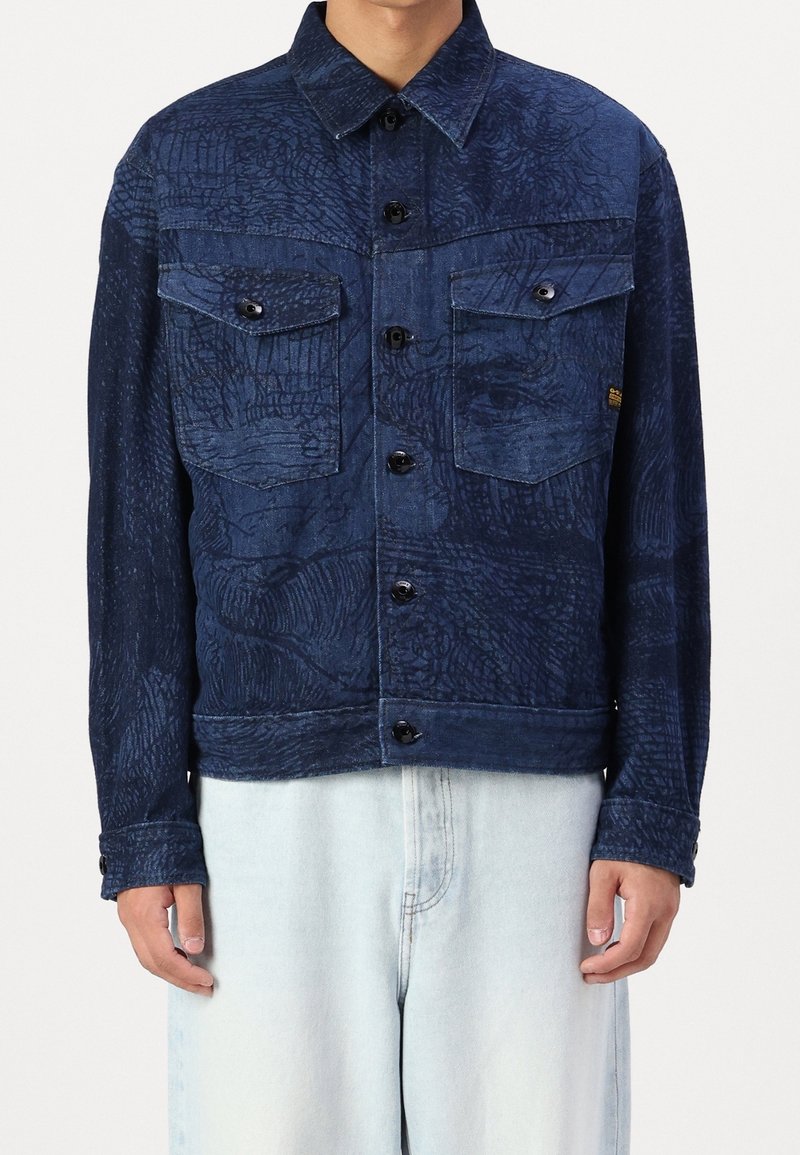 Mörkblå denimjacka med ett strukturerat mönster, med två bröstfickor, knäppning med knappar och rak passform, som matchas med ljusa jeans.