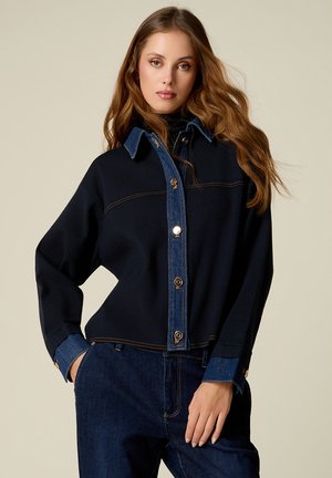 Frau mit langen, welligen Haaren trägt eine schwarze Jacke mit blauem Denimkragen, Manschetten und Knopfleiste, kombiniert mit dunkelblauen Jeans.
