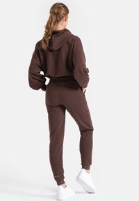 Brązowy, krótki hoodie z oversize'owymi rękawami w komplecie z pasującymi dresami. Posiada sznurki w talii oraz białe akcenty na sneakersach. Miękka tekstura.