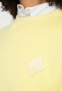 Żółta dzianinowa bluza z teksturowaną, kwadratową naszywką ozdobioną ukośnymi białymi liniami. Prosty krój z okrągłym dekoltem i miękką fakturą.