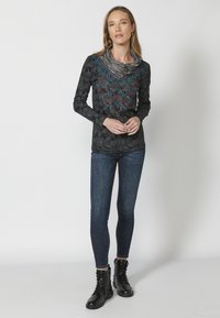 Langärmliges schwarzes Oberteil mit filigraner blauer und roter Stickerei am Ausschnitt, kombiniert mit schmal geschnittenen blauen Jeans und schwarzen Schnürstiefeln.
