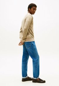 Beige kabelbreisel trui, blauwe rechte jeans, en zwarte instappers. De trui heeft ribgebreide manchetten; de jeans zijn lichtjes opgerold aan de onderkant.