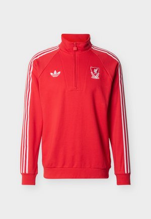 Czerwony sportowy pulower z długim rękawem z białym logo Adidas, herbem Liverpool FC, białymi trzema paskami na rękawach i zamkiem błyskawicznym z przodu przy kołnierzu.