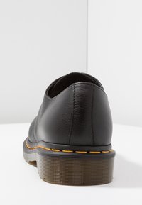 Chaussure en cuir noir avec un talon arrondi, présentant une semelle en caoutchouc côtelé et des coutures orange le long du bord. Texture lisse, design minimaliste.