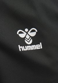 Weißes stilisiertes Hummel-Logo und „hummel“-Text auf strukturiertem schwarzem Stoffhintergrund.