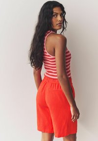 Shorts rossi in lino con vita elastica e tasche laterali, abbinati a una canotta a righe rosa e bianche. Capelli lunghi e mossi neri.