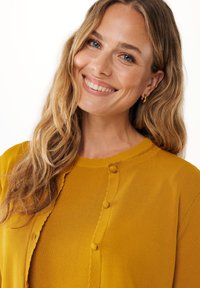 Hořčicově žlutý pletený kardigan s vlněným límcem, texturovanou tkaninou a kulatými knoflíky vpředu. Vlasy upravené do volných vln.