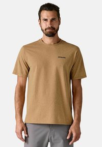 Camiseta marrón de manga corta hecha de tela suave, con cuello redondo y un logo negro de Patagonia en el lado izquierdo del pecho.