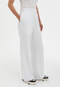 White, wide-leg sweatpants with an elastic waistband, featuring side pockets and a soft, smooth texture. Accented by black sneakers.