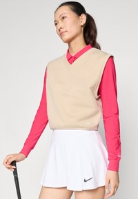 Beige gebreide mouwloze trui over een felroze langemmouwen shirt en een witte geplooide rok, met Nike-logo's op de kledingstukken.