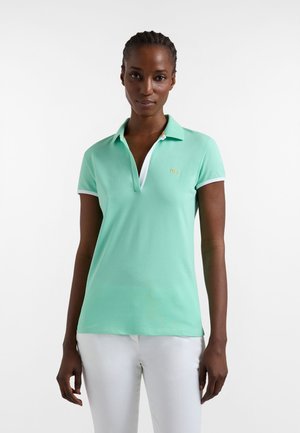 Donna che indossa una polo a maniche corte verde menta con bordi bianchi e pantaloni bianchi, in piedi davanti a uno sfondo bianco uniforme.