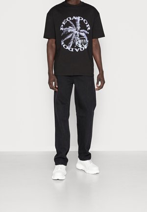 Zwart T-shirt met een grafisch ontwerp van een palmboom en witte letters, gecombineerd met zwarte spijkerbroek en grove witte sneakers.