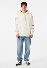 Felpa color crema con tasca frontale, caratterizzata da diverse stampe grafiche colorate sulle maniche. Indossata con jeans azzurri e sneaker nere e bianche.