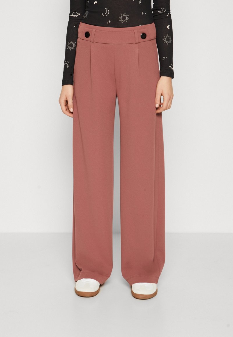 Pantalons larges couleur rose avec une texture lisse, dotés d'une taille haute, de plis et d'accents de boutons noirs à la taille.