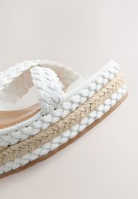 Sandale à bride tressée blanche avec une semelle intermédiaire en jute tissée et une semelle extérieure beige, présentée en gros plan sur un fond clair.