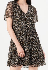 Žena v krátkých černých šatech s hnědými a béžovými abstraktními skvrnami, s výstřihem do V a průhlednými krátkými rukávy, stažené v pase šňůrkou.