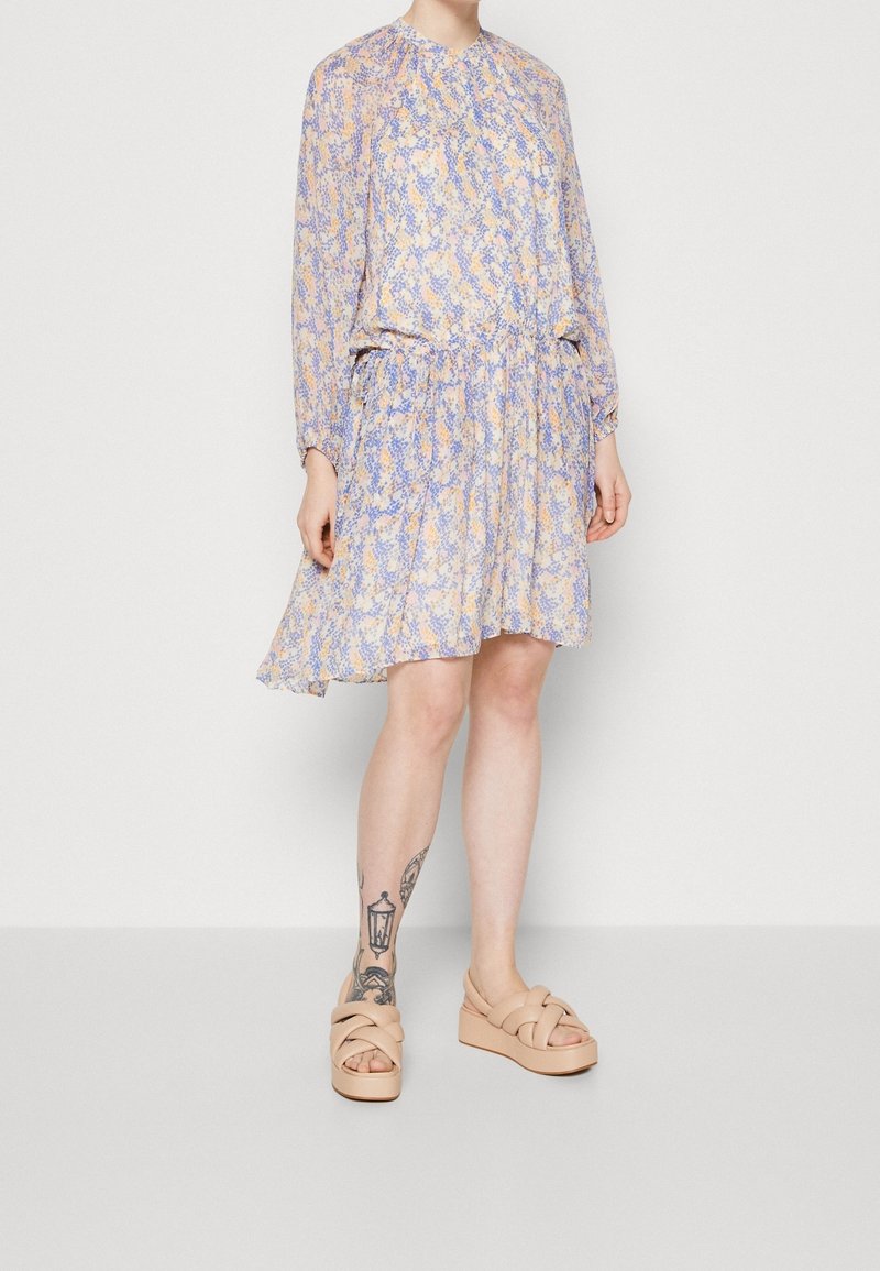 Femme portant une robe à manches longues avec un motif violet et beige, taille froncée, et des sandales à plateforme beiges, montrant un tatouage sur la jambe gauche.