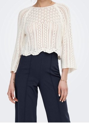 Femme portant un top court en crochet blanc à manches longues et un pantalon noir taille haute à jambes larges, avec peu de bijoux sur les mains.