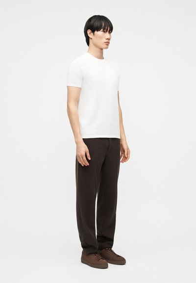 Άνδρας όρθιος, φορώντας ένα απλό λευκό κοντομάνικο Τ-shirt, σκούρο καφέ παντελόνι και καφέ αθλητικά, μπροστά σε ουδέτερο φόντο.
