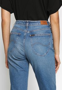 Jean en denim bleu à taille haute, avec une étiquette en cuir marron à l'arrière et deux poches arrière ornées de détails de couture courbes.