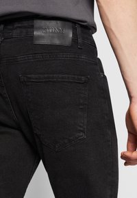 Juodos džinsinės kelnės su odiniu prekės ženklo žyme ant juosmens ir matoma užpakaline kišene. Audinys yra lygus, tekstūruotas.