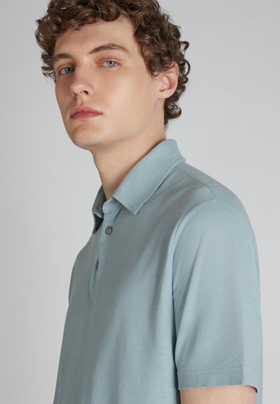 Polo bleu clair en tissu doux, doté d'un col court, d'une patte de boutonnage à trois boutons et d'une texture subtile. Des manches courtes complètent le design.