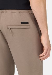 Svijetlosmeđi trenirka od mekog materijala, s patentnim zatvaračem na stražnjem džepu, elastičnom strukom i malom crnom etiketom s natpisom "INTECH."