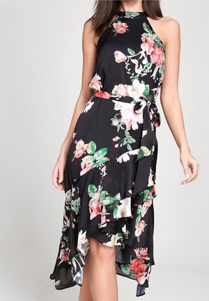 Mujer con vestido negro sin mangas con estampado floral, cuello alto, bajo asimétrico en capas y cinturón atado a la cintura.