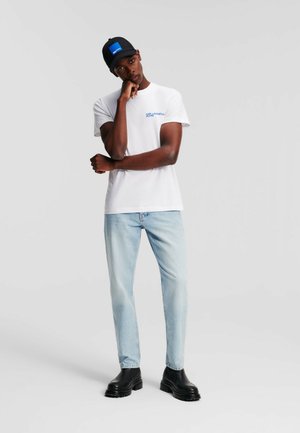 T-shirt blanche avec logo bleu, jean bleu clair, et bottes noires à semelles épaisses ; portant une casquette noire avec un accent carré bleu ; tenue décontractée.