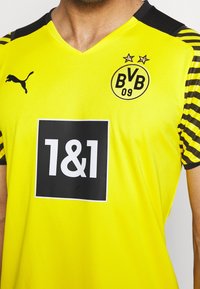 Gul sporttröja med svarta detaljer, med v-ringning, logotypmärken och en stor svart rektangulär sponsorlogo.