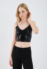Black synthetic cropped top with thin straps, featuring a fitted design and glossy finish, paired with high-waisted black trousers.