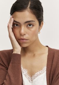 Cardigan maro cu mansete cu nervuri, purtat peste un top alb cu dantelă la margine. Mana persoanei se sprijină pe față, conturând trăsăturile acesteia.