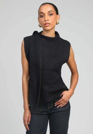 Femme portant des boucles d'oreilles créoles en or, un haut noir sans manches en tricot et un jean foncé, posant devant un fond gris.