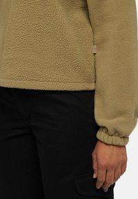 Chaqueta de forro polar en beige, con una superficie texturizada, puños elásticos y una pequeña etiqueta con el logo. Combinada con pantalones cargo negros.