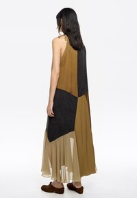 Długa sukienka bez rękawów z kolorowym wzorem w odcieniach oliwkowym, czarnym i beżowym. Wykonana z lekkiego materiału z przezroczystymi akcentami.