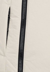 Jasno beżowa kurtka z czarnym zamkiem błyskawicznym, pikowaną teksturą i boczną kieszenią, zaprojektowana w eleganckim, minimalistycznym stylu.