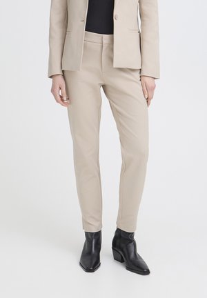 Kvinde iført beige skræddersyede bukser, matchende blazer, sort top og sorte ankelstøvler med hæl, stående på hvid baggrund.