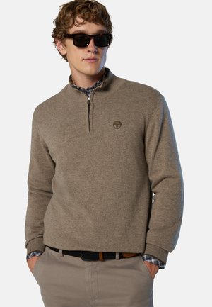 Hombre joven con gafas de sol, suéter beige con media cremallera sobre una camisa a cuadros, con las manos en los bolsillos y fondo neutro.