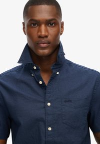 Camisa de botón de manga corta en color azul marino con cuello puntiagudo, que cuenta con un bolsillo en el pecho, un pequeño detalle de logo y botones blancos.