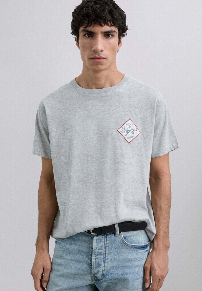 Camiseta de algodón gris con mangas cortas, que presenta un logo de diamante rojo y blanco en el pecho, combinada con jeans de mezclilla azul claro.