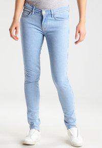 Jeans ajustados de color azul claro hechos de denim, con un diseño de cinco bolsillos, presillas para cinturón y cierre de botón en la parte frontal. Combinados con zapatillas blancas.