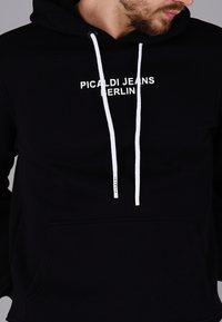 Black hoodie made of soft fabric, featuring a front pocket, white drawstrings, and the text “PICALDI JEANS BERLIN” in white.
